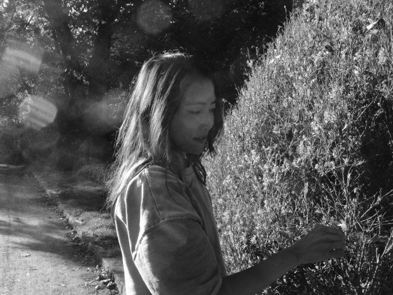 Das Profil einer aus Asien stammenden Frau, Foto in schwarz weiss. Lange schwarze Haare bedecken halb das Gesicht. Im Hintergrund steht ein Gebüsch, dass sich wie eine Aura das Porträt umrahmt. Licht und Schattenspiel.